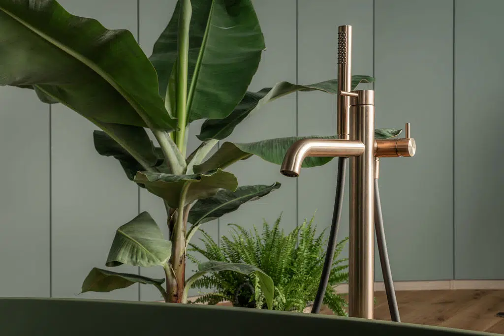 Impressie van een Gessi vloerkraan in de kleur warm bronze. De kraan staat bij een vrijstaand bad in olijfgroen. Achter de kraan zien we een tropische plant.