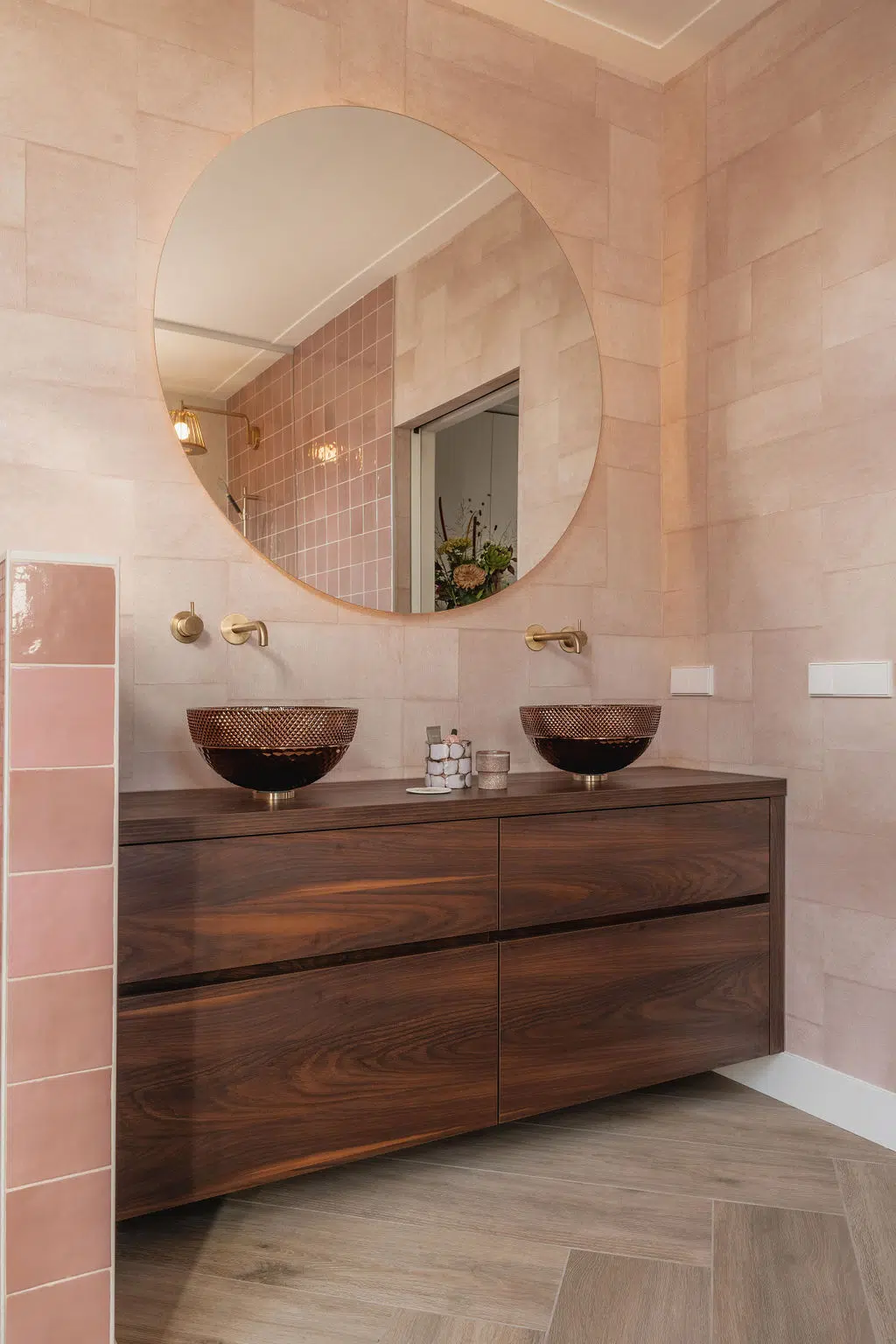 Impressie van een luxe badkamer, ingericht met roze wandtegels, visgraat vloertegels, een ronde badkamerspiegel, en een donker badkamermeubel, glazen waskommen en kranen in geborsteld messing.